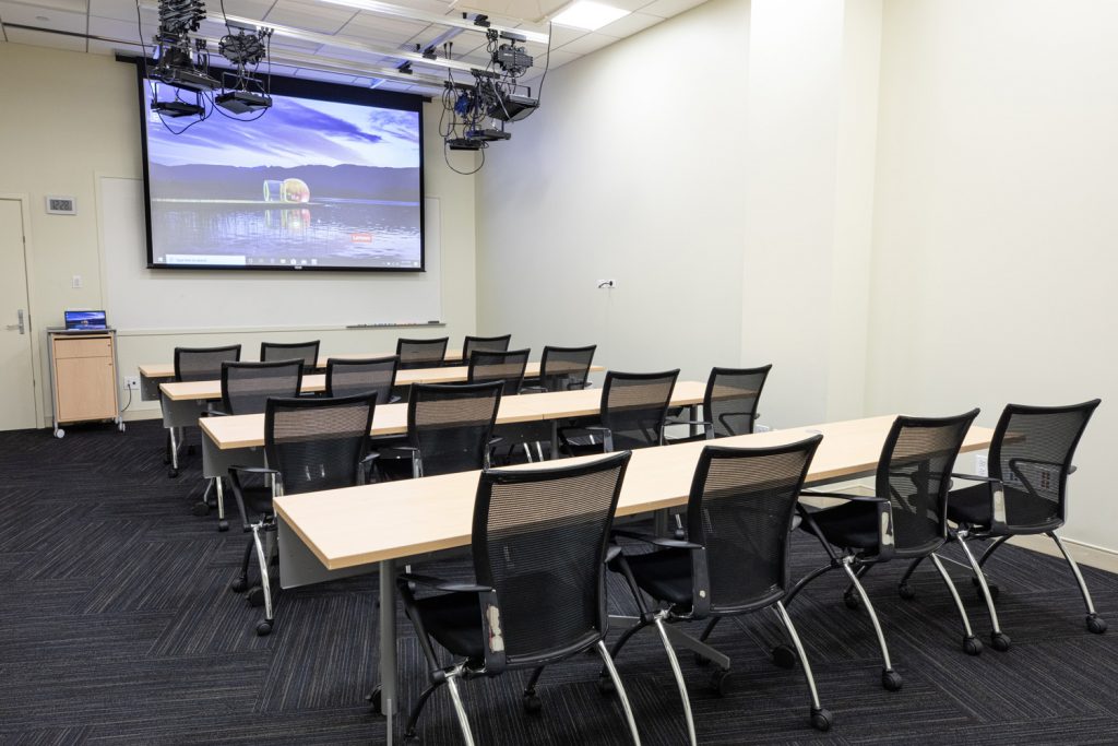 1st Floor Studio Conference Room at One Rotary Place. Photo of a small conference room with tables & chairs, a projector, and screen.
