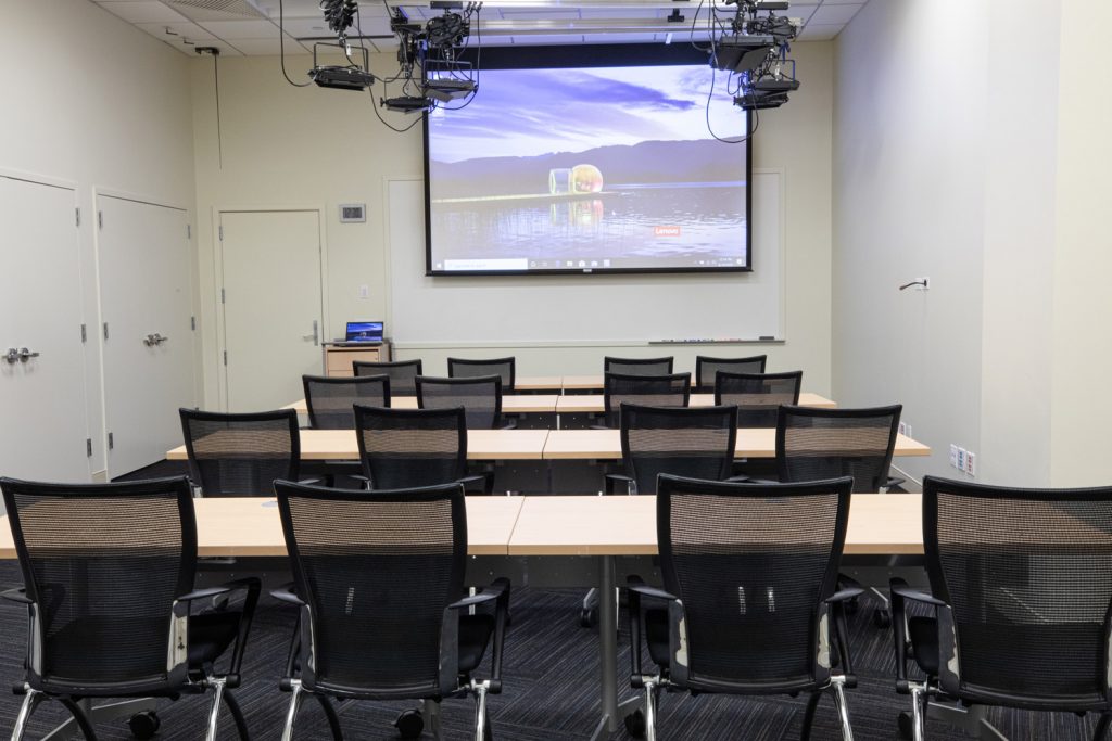 1st Floor Studio Conference Room at One Rotary Place. Photo of a small conference room with tables & chairs, a projector, and screen.