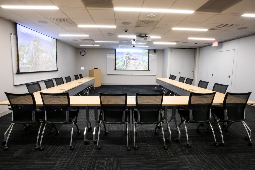 Conference Room 3B at One Rotary Place. Photo of a large conference room with tables & chairs, a podium, multiple projectors & screens, and multiple whiteboards.