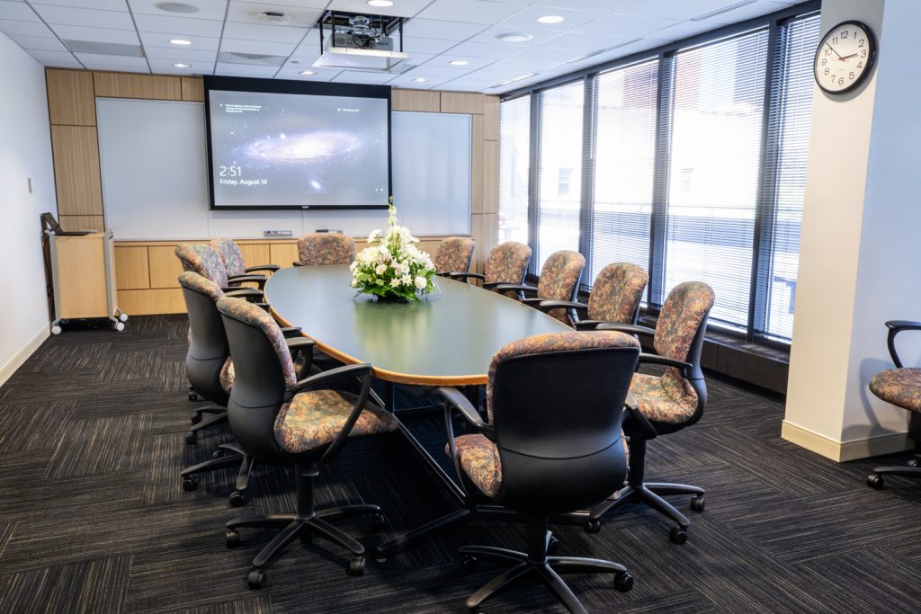 3 East Conference Room at One Rotary Place. Photo of a an executive conference room with a center table, projector & screen, and a whiteboard.