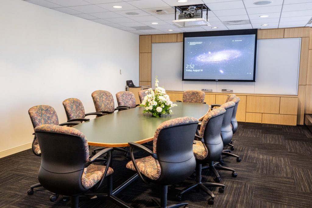 3 East Conference Room at One Rotary Place. Photo of a an executive conference room with a center table, projector & screen, and a whiteboard.